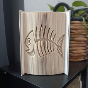 Puede incluir: Una escultura de libro con un diseño de páginas plegadas que crea la silueta de una espina de pescado. Las páginas son de color crema con texto impreso en negro visible, y el libro está de pie con las cubiertas abiertas para mostrar el arte.