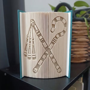 May include: A folded book art sculpture featuring the crossed flail and crook symbols of ancient Egypt. The pages are precisely folded to create the relief design, with the teal-coloured book covers visible on the left and right sides of the sculpture.