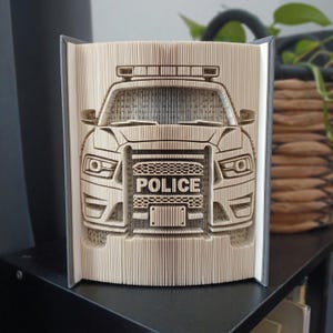 Könnte beinhalten: Eine Buchfaltkunst-Skulptur, die die Vorderansicht eines Polizeiautos darstellt. Die Seiten sind präzise gefaltet, um die Form des Fahrzeugs mit Kühlergrill, Scheinwerfern und Lichtbalken zu bilden. Das Wort POLICE ist auf dem Kühlergrill zu sehen.