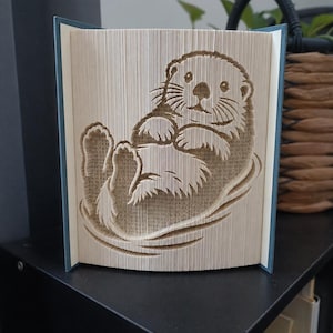 Peut inclure: Une sculpture de livre présentant un design d'art en pages pliées représentant une loutre de mer flottant sur le dos. Les pages sont précisément pliées pour former la silhouette détaillée de l'animal, avec le visage, les pattes et le corps.