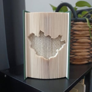 May include: A book with folded pages creating a cutout shape of the map of Iceland. The book has a dark green cover and is standing upright with the pages fanned out to reveal the intricate paper sculpture formed by the precisely folded white paper.