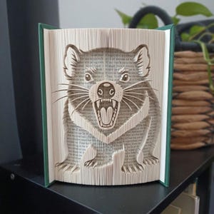 Puede incluir: Una escultura de libro plegado que muestra un demonio de Tasmania con la boca abierta y los dientes al descubierto. Las páginas están dobladas con precisión para crear la imagen tridimensional del animal, sobre las cubiertas verde oscuro.