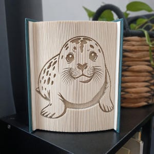 Può includere: Una scultura realizzata con un libro piegato che raffigura la sagoma dettagliata e tridimensionale di una foca maculata. Le pagine sono piegate e incise con precisione per formare l'immagine del muso e del corpo della foca, su una copertina verde ottanio.