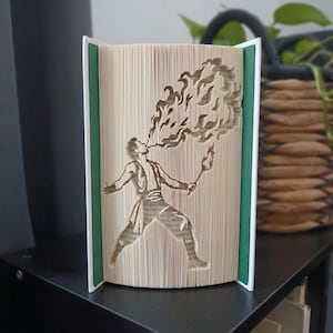 Peut inclure: Une sculpture en livre plié représentant une silhouette crachant du feu. Les pages sont minutieusement pliées pour former le contour d'une personne tenant une torche et exhalant des flammes. Le livre a des couvertures blanches et des panneaux intérieurs verts.