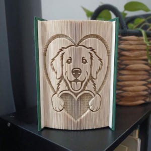 Puede incluir: Una escultura de libro plegado que presenta la cara de un perro centrada dentro de una forma de corazón. Las páginas están dobladas con precisión para crear la imagen en relieve de un canino con la lengua fuera, enmarcado por un corazón grande.