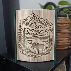 Könnte beinhalten: Eine Buchfaltkunst-Skulptur, die durch präzises Falten der Seiten ein Gebirge, Tannen und die Silhouette eines Elchs darstellt. Das Buch hat einen dunkelgrünen Einband und die Innenseiten zeigen den durch die Faltungen sichtbaren Text.