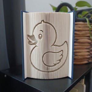 May include: A book art sculpture featuring a duck silhouette created by precisely folding the pages of a book. The book has a dark blue cover, and the interior pages are arranged to form the distinct shape of a duck with a visible eye and wing.