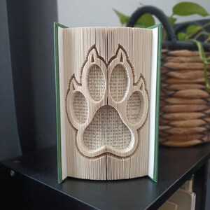 Può includere: Una scultura realizzata con un libro piegato che presenta un design dettagliato a forma di impronta di zampa di animale, creato piegando con precisione le singole pagine di un libro con copertina rigida. Le pagine color crema formano una zampa.