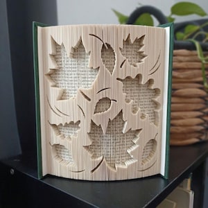 Peut inclure: Une sculpture en livre plié présentant des découpes complexes de diverses formes de feuilles. Les pages sont précisément pliées et marquées pour créer un motif tridimensionnel de silhouettes d'érables et d'autres feuillages sur le texte.