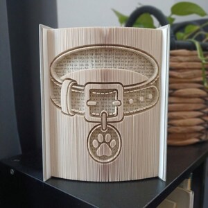 Könnte beinhalten: Eine Buchfaltkunst-Skulptur mit einem detaillierten Design eines Tierhalsbandes mit Schnalle und einem runden Anhänger, der einen Pfotenabdruck zeigt. Die Skulptur entsteht durch präzises Falten der Buchseiten zu einem dreidimensionalen Bild.