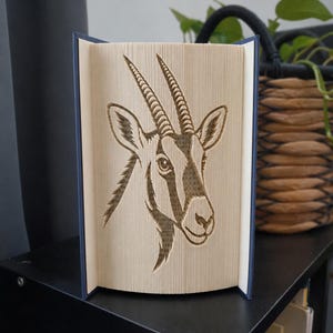 May include: A book sculpture featuring a folded page art design of an oryx head. The pages are precisely creased to form the detailed silhouette of the animal, including its long, straight horns, pointed ears, and facial markings in beige paper.