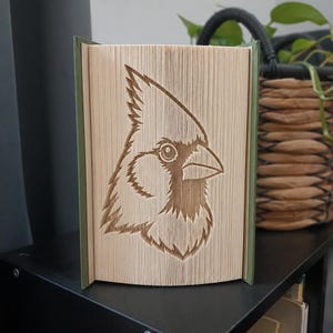 Könnte beinhalten: Eine Buchfaltkunst-Skulptur, die das detaillierte Profil eines Kardinalvogels zeigt. Die Seiten sind präzise gefaltet und geknickt, um das Bild des Vogelkopfes mit spitzem Kamm, Auge und Schnabel vor einem grünen Einband zu erzeugen.
