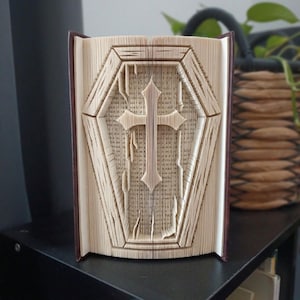 Peut inclure: Une sculpture en livre plié représentant un cercueil avec une croix centrale. Les pages sont précisément pliées et marquées pour créer un relief tridimensionnel en forme de cercueil avec une croix, mettant en valeur le texte imprimé.