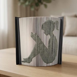 Peut inclure: Sculpture de livre unique représentant une silhouette d'une personne lisant. Les pages sont pliées pour créer l'image, avec une tranche et des bords noirs. Le livre est posé sur une surface en bois clair.