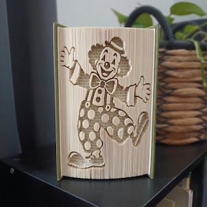 May include: A folded book art sculpture featuring a clown design created by precisely folding the pages of a hardback book. The clown figure is depicted with a hat, bowtie, suspenders, and polka-dot trousers, standing in a cheerful pose.