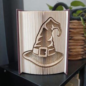 Può includere: Una scultura realizzata con un libro piegato che raffigura un cappello da strega, creata piegando con precisione le singole pagine di un libro con copertina rigida. Le pagine sono aperte a ventaglio per rivelare la sagoma di un cappello a punta.