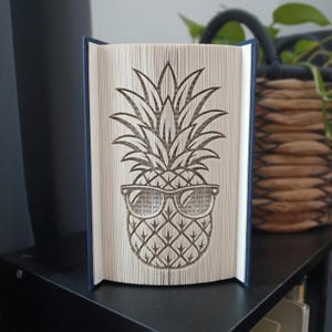 Könnte beinhalten: Eine Buchfaltkunst-Skulptur mit einem Ananas-Motiv und einer Sonnenbrille. Die Seiten sind präzise gefaltet, um die Form der Ananas zu erzeugen, wobei der Text des Buches auf den Seitenrändern sichtbar ist. Das Buch hat einen dunkelblauen Einband.