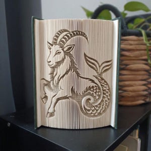 Puede incluir: Una escultura de libro plegado que representa el signo zodiacal Capricornio. Las páginas están dobladas con precisión para crear una imagen tridimensional de una cabra marina con cuernos curvos, cabeza de cabra y cola de pez con escamas.