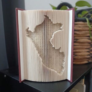 Könnte beinhalten: Ein Buch mit gefalteten Seiten, die eine dreidimensionale Silhouette der Landkarte von Peru bilden. Das Buch hat einen dunkelroten Einband und die cremefarbenen Seiten zeigen gedruckten schwarzen Text, der die Form des Landes nachbildet.