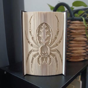 May include: A folded book sculpture featuring a detailed spider design created by precisely folding the individual pages of a hardcover book. The spider is centered on the page edges, displaying eight legs and a patterned abdomen in beige.