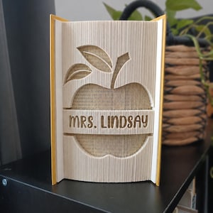 Op de afbeelding: Een handgemaakte boeksculptuur met een appelontwerp en de naam "MRS. LINDSAY" uitgesneden in de pagina's. Het boek heeft gele randen en staat op een zwarte plank. Het appelontwerp is lichtbruin.