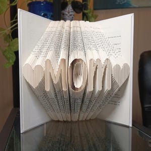 May include: A decorative book sculpture spelling "MOM" from folded pages. The letters are cut out, and the pages are fanned to create a 3D effect. Two heart shapes flank the word. The book is open and displayed on a glass surface, a unique gift idea.