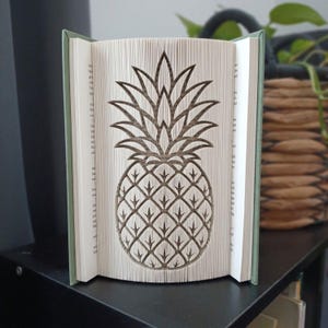 Peut inclure: Une sculpture en livre plié représentant un ananas, réalisée en pliant avec précision les pages individuelles d'un livre à couverture rigide. Le livre possède une couverture vert sauge et des pages blanches, formant la silhouette de l'ananas.