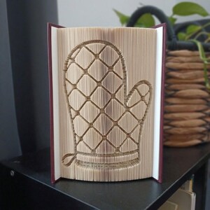 May include: A folded book art sculpture featuring a quilted oven mitt design created by precisely folding the individual pages of a hardcover book. The beige paper edges form a clear diamond pattern with a thumb section and a small hanging loop.