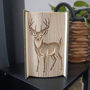 May include: A folded book art sculpture featuring a detailed silhouette of a standing deer with antlers. The pages are precisely creased and folded to create the three-dimensional image of the animal, showcasing the text on the paper edges.