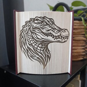 Puede incluir: Una escultura de libro plegado que presenta el perfil detallado de una cabeza de caimán. Las páginas están dobladas con precisión para crear una silueta oscura y de alto contraste del reptil, mostrando sus escamas, dientes y ojo.