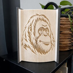 Puede incluir: Una escultura de libro plegado que presenta un diseño detallado de una cara de orangután, creado doblando con precisión las páginas individuales de un libro. Los bordes de papel beige claro forman el retrato, con las cubiertas oscuras visibles.
