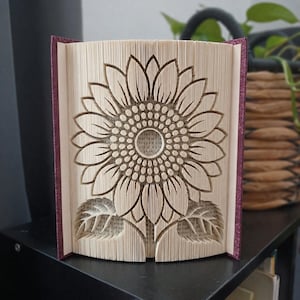 May include: A folded book art sculpture featuring a detailed sunflower design created by precisely folding the pages of a book. The sculpture has a dark purple cover and displays a large, intricate floral pattern with leaves at the base.