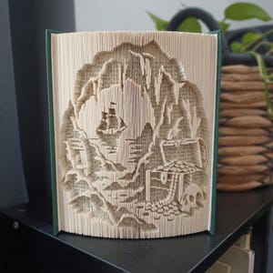 Könnte beinhalten: Eine Buchfaltkunst-Skulptur mit einer detaillierten Szene eines Segelschiffs auf dem Wasser, einer Felsklippe und einem kleinen Gebäude mit einem Totenkopf an der Basis. Die Seiten sind präzise gefaltet, um ein dreidimensionales Reliefbild zu erzeugen.