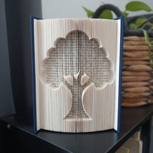 Op de afbeelding: Een boeksculptuur met gevouwen pagina's die het silhouet van een boom vormen. Het boek heeft een donkerblauwe kaft en de binnenpagina's zijn crèmekleurig met zichtbare zwarte gedrukte tekst. Het boomontwerp is gecentreerd in het boek.