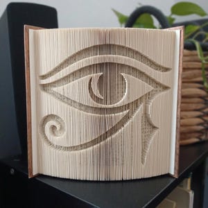 Puede incluir: Una escultura de libro que presenta el Ojo de Horus, creada doblando con precisión las páginas individuales de un libro de tapa dura. Los bordes de papel color crema están manipulados para formar el distintivo símbolo egipcio.