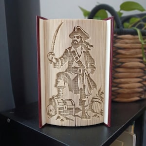 May include: A folded book art sculpture featuring a detailed silhouette of a pirate standing with a sword raised, positioned next to a treasure chest and a skull. The pages are precisely folded to create the image, with a dark red hardback cover.