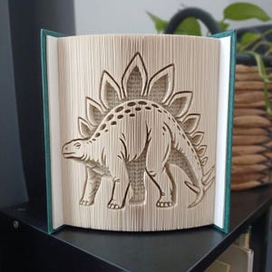 Peut inclure: Une sculpture de livre présentant un design d'art en pages pliées représentant un dinosaure Stégosaure. Les pages sont minutieusement pliées pour créer la silhouette du dinosaure avec ses plaques dorsales, dans une couverture de livre rigide bleu sarcelle.