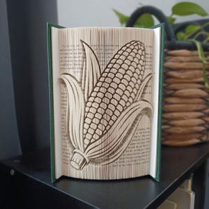 Op de afbeelding: Een boeksculptuur met een gevouwen paginadesign dat een driedimensionale afbeelding van een maïskolf met bladeren creëert. De pagina's zijn crèmekleurig met zichtbare zwarte gedrukte tekst en het boek heeft een donkergroene kaft.