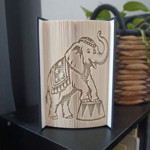 Op de afbeelding: Een boeksculptuur met een gevouwen paginadesign van een olifant die op een circusvoetstuk staat. De olifant is afgebeeld met een decoratieve deken versierd met sterren en kwastjes, met de slurf omhoog gericht, gemaakt van boekpagina's.