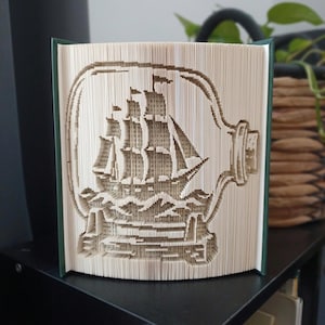 Op de afbeelding: Een gevouwen boekkunstsculptuur met een driedimensionaal ontwerp van een zeilschip in een glazen fles. De pagina's zijn nauwkeurig gevouwen en gesneden om het silhouet van een groot schip op golven te vormen, ingesloten in de omtrek van een fles.