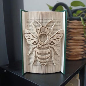 May include: A book sculpture featuring a bee design created by folding the pages of a hardback book. The pages are cream-coloured with black printed text, and the bee shape is formed by the precise folding of the paper edges to create a 3D effect.