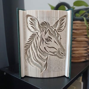 May include: A book sculpture featuring a folded page art design of a deer head in profile. The pages are intricately creased to form the shape of the animal, with the dark green book covers visible on the left and right sides of the open book.