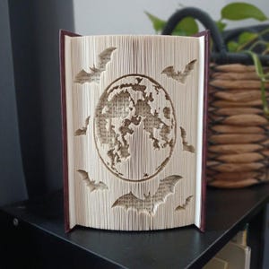 Peut inclure: Une sculpture en livre plié présentant un motif de globe circulaire au centre, entouré de plusieurs silhouettes de chauves-souris découpées dans les pages. Le livre a une couverture bordeaux foncé et les pages sont crème avec du texte imprimé.