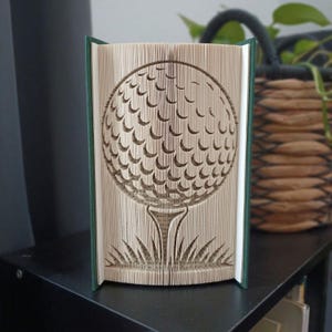 Peut inclure: Une sculpture de livre avec des pages pliées formant la silhouette d'une balle de golf sur un tee, au-dessus d'un brin d'herbe. Le livre possède une couverture vert foncé et est présenté en position ouverte pour révéler cet art du papier.