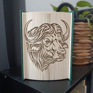 May include: A folded book art sculpture featuring a detailed bull head design created by precisely folding the pages of a hardback book. The book has dark green covers and the pages are arranged to form the intricate silhouette of a bull.