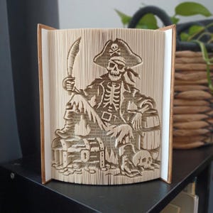 Op de afbeelding: Een gevouwen boekkunstsculptuur met een gedetailleerd piratenskeletontwerp. Het skelet draagt een hoed met een schedel, houdt een gebogen zwaard vast en zit naast een schatkist en een losse schedel, allemaal gevormd uit de boekpagina's.
