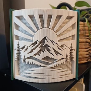 May include: A book sculpture featuring folded pages that create a three-dimensional landscape design. The artwork depicts a sun rising over a mountain range with pine trees on the sides and a reflective water surface at the base of the mountains.