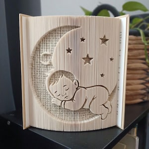 Könnte beinhalten: Eine Buchfaltkunst-Skulptur, die ein schlafendes Baby innerhalb einer Mondsichelform zeigt, umgeben von mehreren kleinen Sternausschnitten. Die Buchseiten sind präzise gefaltet, um das dreidimensionale Bild des Säuglings zu erzeugen.