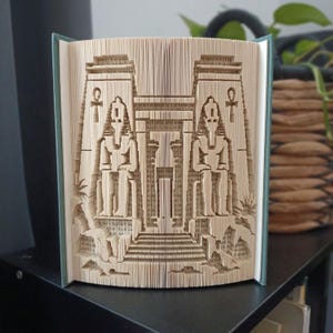 May include: A folded book art sculpture featuring an intricate design of an ancient Egyptian temple entrance. The pages are precisely folded to reveal two standing figures flanking a central doorway, with an ankh symbol visible on each side pillar.