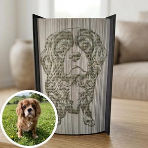 Könnte beinhalten: Dekorative Buchskulptur mit einem detaillierten Bild eines Cavalier King Charles Spaniel Hundes. Das Porträt des Hundes wird durch Falten der Buchseiten erstellt. Ein kleines, rundes Foto des Hundes befindet sich in der unteren linken Ecke.